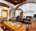 Living room with exposed wooden beams, terracotta tiles, fireplace, sofa, coffee table, rocking chair, bookshelf, and television.