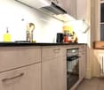 Modern kitchen with light-colored cabinets, stainless steel handles, and a built-in oven. Countertop features a bottle of olive oil, dish soap, and a basket of kitchen utensils. A wooden cutting board with knives and a black kettle are also visible.