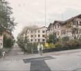 Residential street lined with multi-story houses featuring pitched roofs and varied facades, surrounded by trees and greenery. People are walking along the sidewalk.