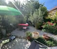 gravel patio with table and chairs, green umbrella, small lounge chair, several potted plants, greenery