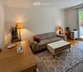 Bright living room with a grey sofa, coffee table, dining table, and a desk area near a large sliding glass door leading to a balcony.