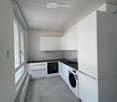 Modern, minimalist kitchen with white cabinets, black countertops, and integrated appliances including a refrigerator, oven, and washing machine. The space features tiled flooring and large windows allowing natural light.