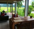 Bright living room with large glass doors opening to a green outdoor area, featuring a black leather sofa with colorful cushions, a wooden dining table with placemats and a decorative crown centerpiece, and a colorful striped chair.