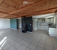 Open-plan space with a tiled floor, exposed wooden ceiling beams, and a modern kitchen area featuring white cabinets, a refrigerator, and a stove. A black wood-burning stove is positioned near the center.
