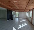 Bright, open room with exposed wooden beams on the ceiling, tiled floor, and large windows allowing natural light to flood the space. The room is completely empty, with no furniture or decorations.
