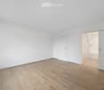 Empty room with light wooden flooring and white walls, featuring a closed door and an open doorway leading to a bathroom.