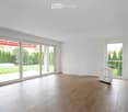 Empty room with wooden flooring, white walls, large sliding glass doors leading to a garden, and a small white decorative stand in the corner.