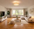 Modern living room with light-colored wooden flooring, large windows, a sectional sofa, coffee table, ottoman, side table, floor lamp, TV stand, and several potted plants.
