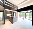 Modern kitchen with grey cabinetry, a central island, and a dining area with a round table and chairs, connected to a spacious living area with large windows and a stone pillar.