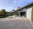 Modern house exterior with large glass sliding doors, minimalist design, and a landscaped garden with a view of a lake.