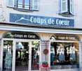 Facade of a clothing store named "Coups de Coeur" with large glass windows displaying mannequins and clothing, a small red table with a potted plant, and a hanging decorative ball.