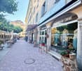 A charming pedestrian street lined with historic buildings, featuring a shop named "Coups de Coeur" with a display window, outdoor seating, and potted plants.