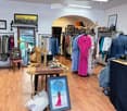 A well-lit clothing boutique with wooden flooring, clothing racks, a wooden table with accessories, and various decorative items including picture frames and a mirror.