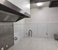 Empty room with white tiled floor, stainless steel walls, and a large exhaust hood. No furniture or equipment present.