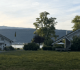 Lush green lawn with trees, bushes, and a trampoline, with houses and a distant mountain range in the background.