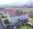 Aerial view of a modern two-story house with a red-tiled roof, white exterior, and a visible garage. The house is surrounded by a well-maintained lawn and is part of a residential neighborhood with other houses and open fields in the background.