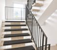 Modern staircase with beige stone steps and black carpet runners, featuring a sleek metal railing and a window at the top landing.