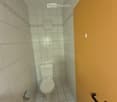 Small bathroom with white tiled walls and floor, a toilet, and a toilet paper holder on the left side. The door has a black handle.