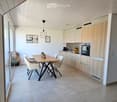 Modern kitchen with light wood cabinets, stainless steel oven, central island, dining table with four chairs, pendant lighting, and decorative wall art.