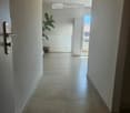 A clean, tiled hallway with a door on the left and an open doorway leading to a balcony area with a plant and outdoor furniture visible.