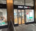 Store with glass entrance, display windows, white shelves, shoes, and various items for sale.