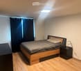 A modern bedroom with a wooden bed, nightstand, lamp, and wall-mounted TV. The room features wooden flooring and dark curtains on the window.