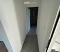 A narrow hallway with white walls and tiled floor, leading to a small shower area with a showerhead and controls.