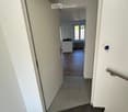 A modern hallway with tiled flooring, white walls, and a staircase leading to an open room with wooden flooring and large windows.