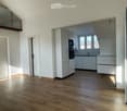 Bright, unfurnished room with wooden flooring, white walls, and large windows. Features a modern kitchen area with white cabinets and stainless steel appliances visible in the background.