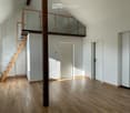 Bright, open loft-style room with wooden flooring, white walls, and a wooden staircase leading to an upper level with glass railings.