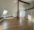 Bright, open attic space with wooden flooring, white walls, exposed wooden beams, skylight window, and a small balcony area with glass railing.