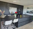 Modern kitchen with dark grey cabinets, a black island, white chairs, a vase with dried flowers, and a large painting on the wall.