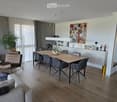 Modern living and dining area with a grey sofa, leather armchair, wooden dining table, grey chairs, large mirror, and decorative plants.