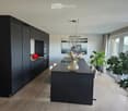 Modern kitchen with dark cabinets, a long black counter, and a vase with dried flowers on the counter. There are chairs around the counter, a large window with a view, and several potted plants near the window.