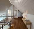 Modern hallway with wooden flooring, slanted wooden ceiling, glass railing, small table with lamp and books, and a mirror on the wall.
