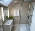 Modern bathroom with a white sink, glass-enclosed shower, and two white towels hanging on the wall. The room features light-colored tiles and a skylight window.