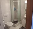 Modern bathroom with white tiled walls and floor, featuring a walk-in shower with glass enclosure, toilet, sink with mirror, and small cabinet.