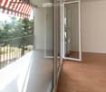 Balcony with wooden floor, glass sliding doors, metal railing, and striped awning. The view shows greenery and a neighboring building.