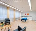 Modern, well-lit room with light wooden flooring, white walls, and large windows covered with sheer curtains. Features include a massage table, two black leather chairs, small wooden tables, a potted plant, a tall floor lamp, and a white cabinet with a co