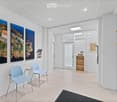 Modern, minimalist waiting area with light blue chairs, white walls, large artwork, and a small wooden shelf holding items.