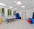 A well-lit therapy room with large windows, a treatment table, exercise balls, a ladder, and a drawer unit.