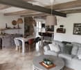 Open-plan living and dining area with exposed wooden beams, a fireplace, a grey sofa, a coffee table, and a dining table with chairs.