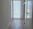 A modern, minimalist hallway with light wooden flooring, white walls, and recessed ceiling lighting. Features a large window with sheer white curtains and several doors along the sides.