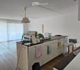 A modern kitchen with white cabinets, a central island, and a sink. There is a sofa in the background, a curtain covering a large window, and various kitchen items on the counter.