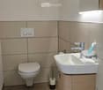 Modern bathroom with a white toilet, a sink with a modern faucet, a toothbrush holder, and a mirror with built-in lighting. The walls are tiled in a light beige color, and the floor is tiled in a darker beige.