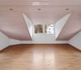 Empty room with wooden flooring and white walls, featuring a slanted wooden ceiling and large windows offering a view of the outside.