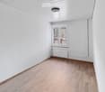 Empty room with light wooden flooring, white walls, and a single window with a view of an outdoor area. The room has a white radiator and a ceiling light fixture.