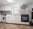 Modern kitchen with white cabinets, a sink, an oven, and a washing machine. The space features wooden flooring and a window above the sink.