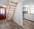 A narrow hallway with a wooden staircase leading upwards, a wooden front door, and a view into a modern kitchen area with white cabinets and a washing machine.
