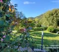 A well-maintained garden with a variety of flowering plants, including roses, and a neatly trimmed lawn. The background features dense greenery and a hill under a clear blue sky.